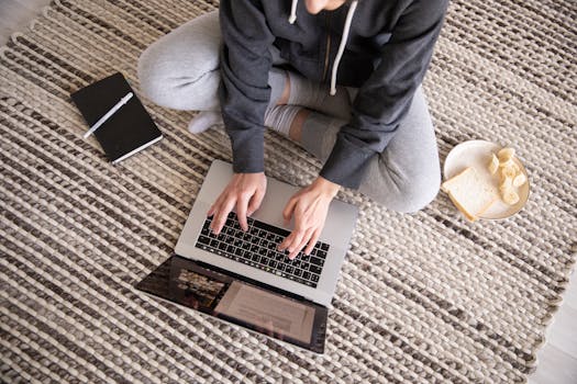 Woman in casual wear working remotely on a laptop, with a notebook and snack nearby.
